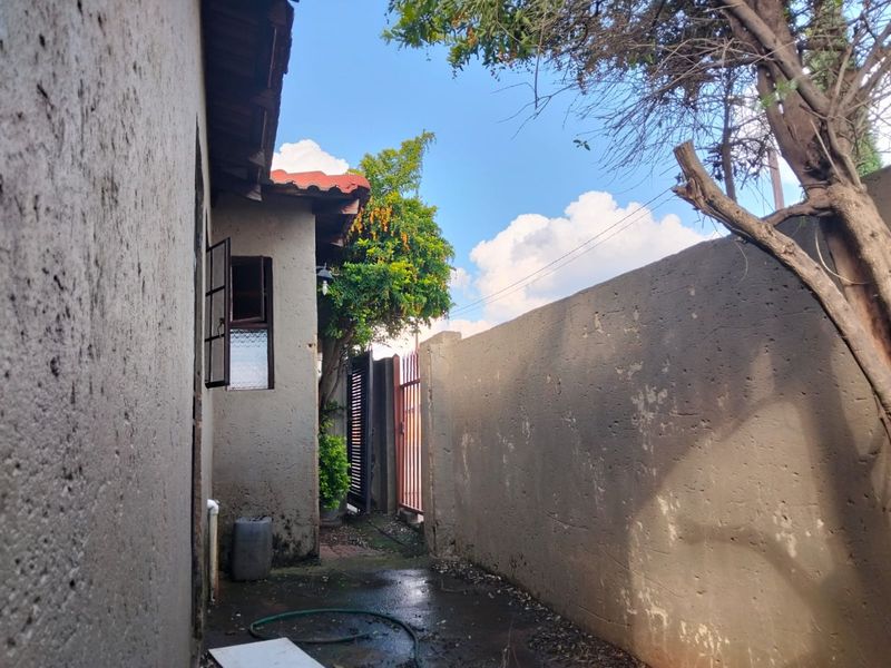 A narrow side passage of the property featuring a boundary wall, metal gate, and exterior wall access with visible signs of weathering.
