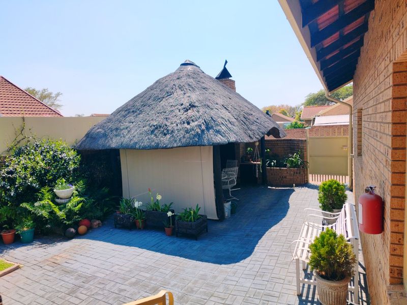 A well-maintained rear courtyard featuring a traditional thatched roof lapa, brick paving, and established garden borders.