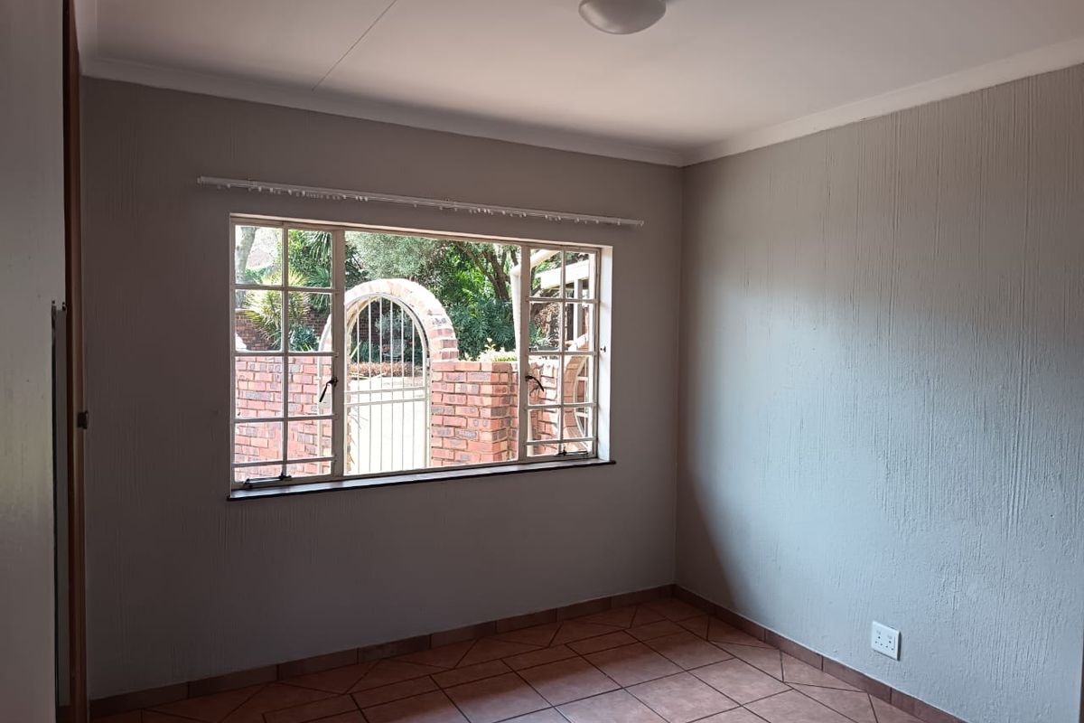 An empty bedroom featuring tiled flooring, textured walls, and a steel-framed window overlooking a garden area.