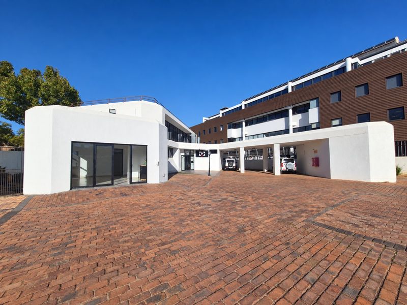 Front exterior view of a commercial property featuring a paved courtyard, covered parking bays, and modern white building facade with glass entrance doors.