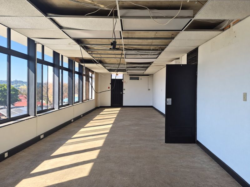 An empty commercial office space featuring large windows, carpeted flooring, and a suspended ceiling requiring repair and maintenance.
