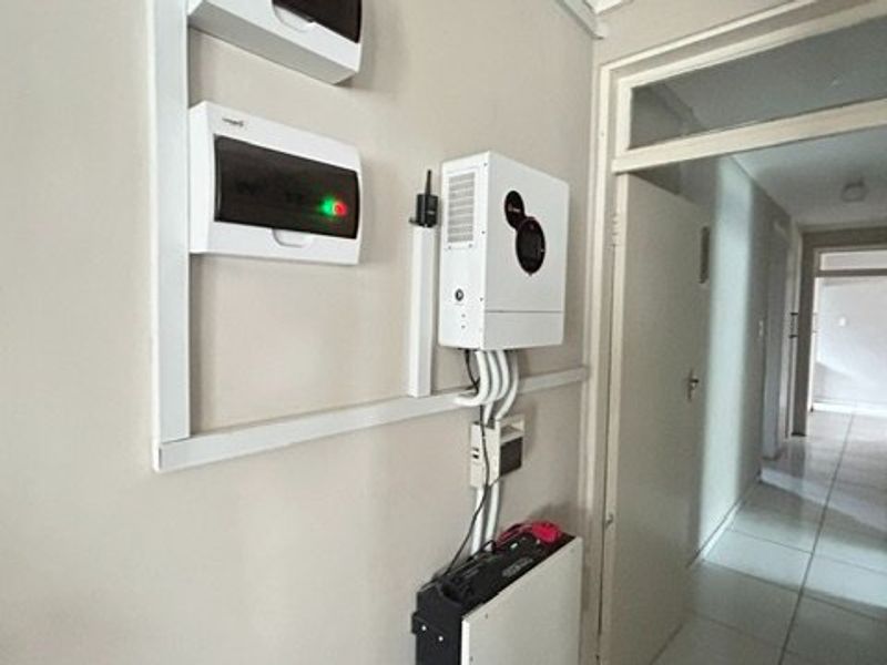 Utility area featuring a wall-mounted solar inverter, lithium battery storage system, and electrical distribution board, leading into a tiled hallway.