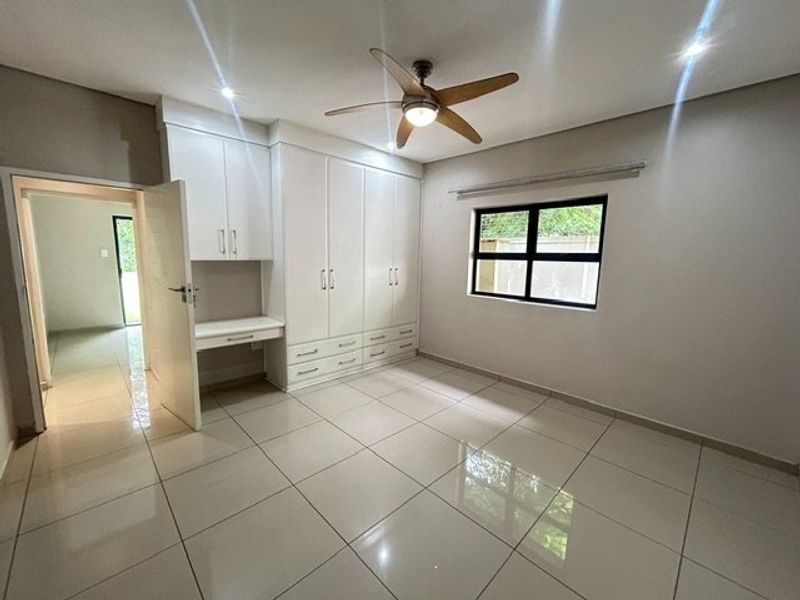 A bright, modern bedroom featuring large-format floor tiles, extensive built-in cabinetry with an integrated desk, and a ceiling fan.