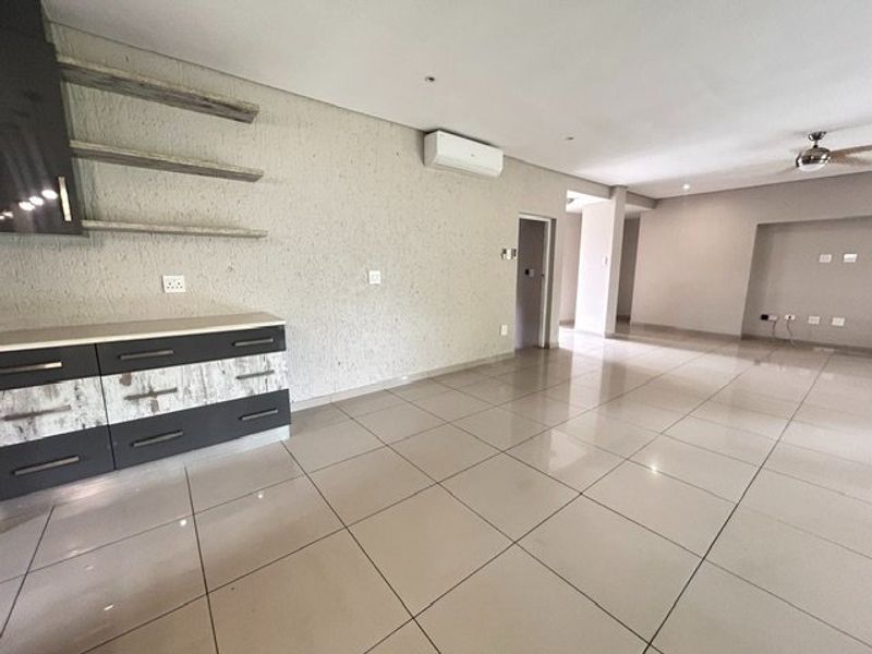 Spacious open-plan living area featuring neutral tiled flooring, built-in cabinetry with floating shelves, and a wall-mounted air conditioning unit.