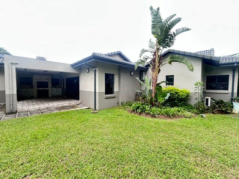 Rear view of the property featuring a covered patio with a built-in braai, overlooking a well-maintained lawn and landscaped garden area.