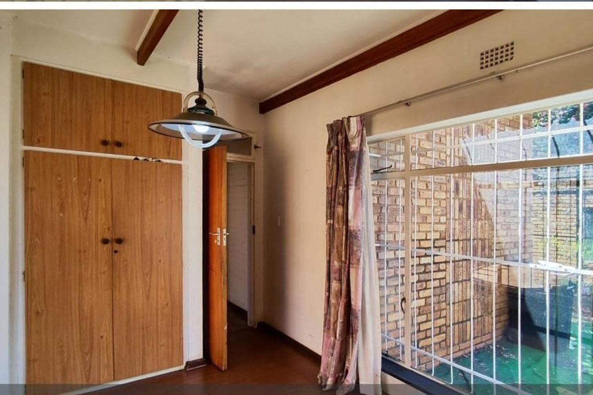 A bedroom featuring built-in wooden cupboards, exposed ceiling beams, and large windows with security bars, offering a functional space ready for modernization.