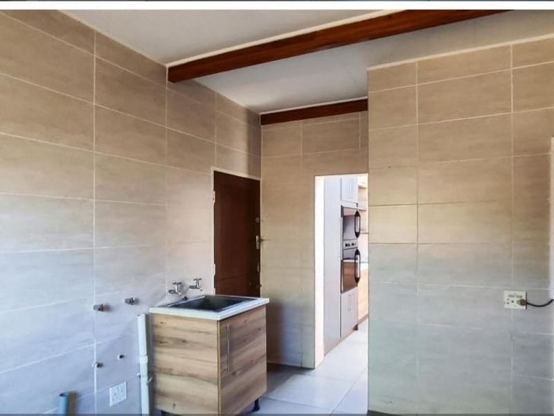 A functional laundry area featuring full-height wall tiling, a stainless steel sink, and plumbing connections, with a view through to an adjacent kitchen space.