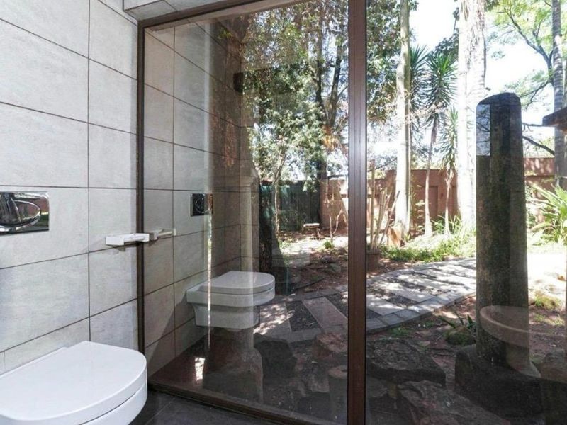 A modern bathroom featuring a wall-mounted toilet, large glass window overlooking a private garden, and neutral tiled finishes.