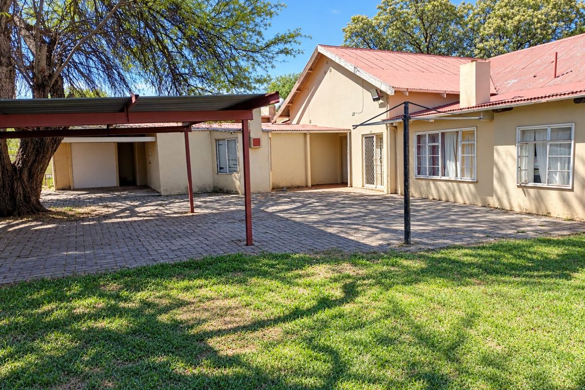 Rear view of the property featuring a paved courtyard, attached carport, and established garden area with mature trees.