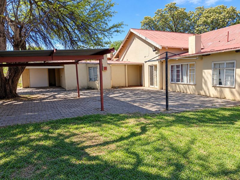 Rear view of the property featuring a paved courtyard, attached carport, and established garden area with mature trees.