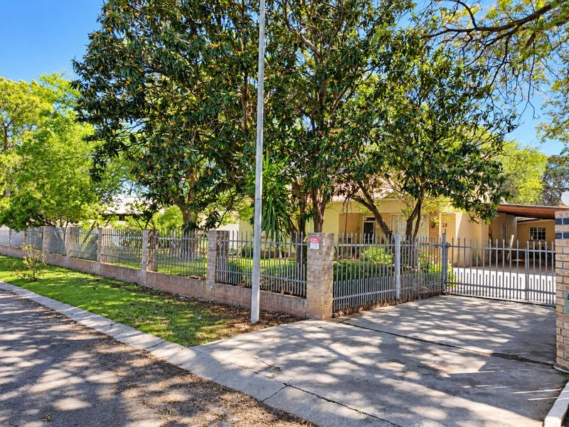Front view of the property featuring a concrete driveway, steel gate, and secure perimeter fencing with established trees.