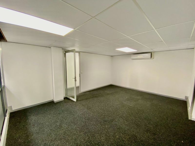 A clean, empty office space featuring a suspended ceiling with recessed lighting, carpeted flooring, and a wall-mounted split-system air conditioning unit.