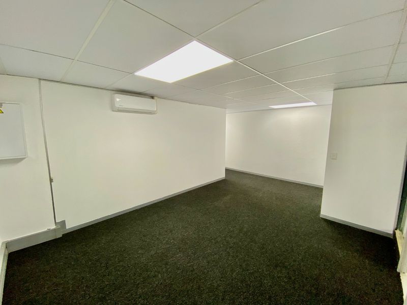 A clean, empty commercial office space featuring carpeted flooring, a suspended ceiling with recessed lighting, and a wall-mounted air conditioning unit.