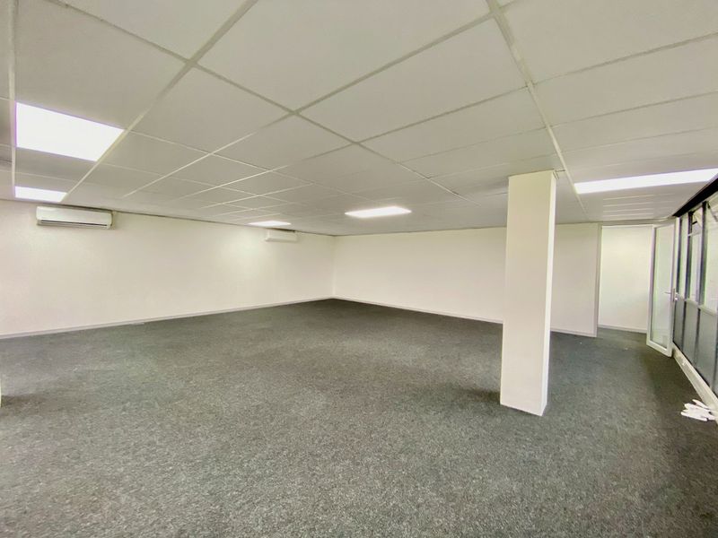 A spacious, open-plan commercial office area featuring a suspended ceiling, recessed lighting, carpeted flooring, and wall-mounted air conditioning units.