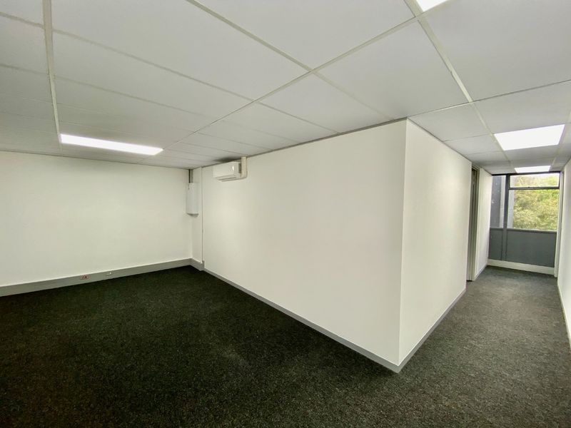 A clean, vacant office space featuring a suspended ceiling, recessed lighting, split-system air conditioning, and dark carpet flooring with access to a windowed area.