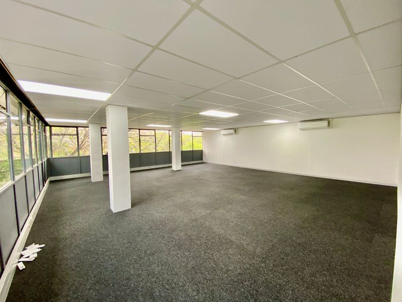 A spacious, open-plan commercial office area featuring a suspended ceiling, recessed lighting, split-system air conditioning, and large windows providing natural light.