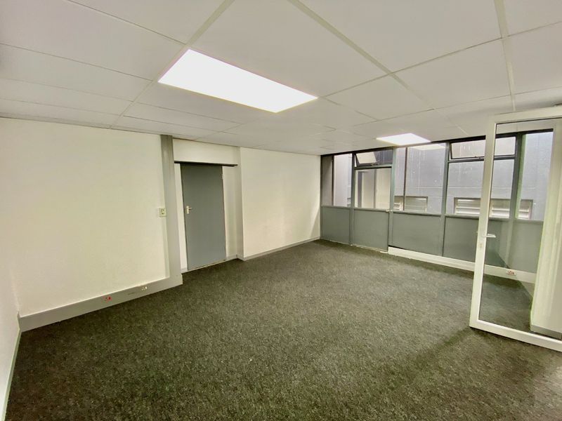 A vacant, well-lit office space featuring a suspended ceiling, carpeted flooring, and an internal glass partition wall providing natural light.