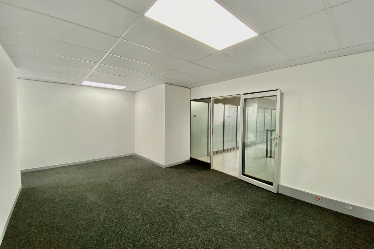 A clean, empty commercial office space featuring grey carpeting, a suspended ceiling with recessed lighting, and a glass partition entrance.