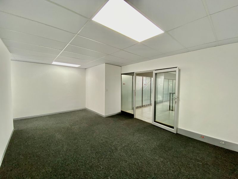 A clean, empty commercial office space featuring grey carpeting, a suspended ceiling with recessed lighting, and a glass partition entrance.