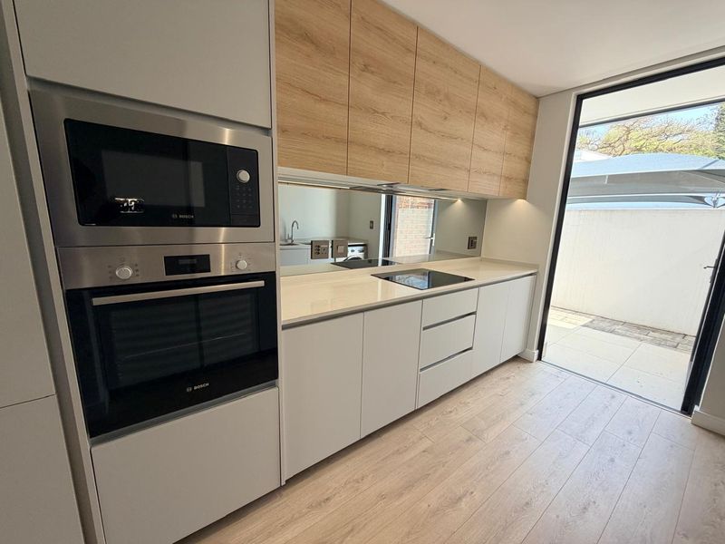 Modern kitchen featuring light wood-tone cabinetry, stone countertops, integrated Bosch appliances, and a mirror splashback, with direct access to an outdoor patio area.