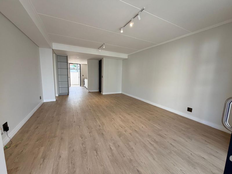 A bright, spacious open-plan living area featuring modern laminate flooring, neutral walls, and adjustable track lighting, leading towards a kitchen area.