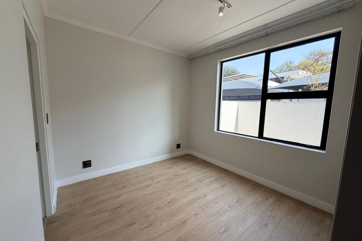 A bright, empty bedroom featuring modern laminate flooring, neutral walls, and a large black-framed window with track lighting.