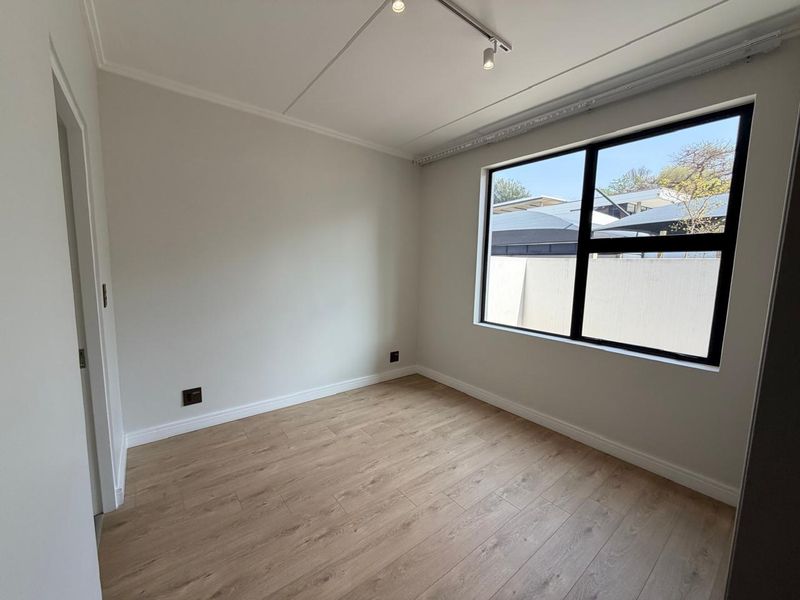 A bright, empty bedroom featuring modern laminate flooring, neutral walls, and a large black-framed window with track lighting.