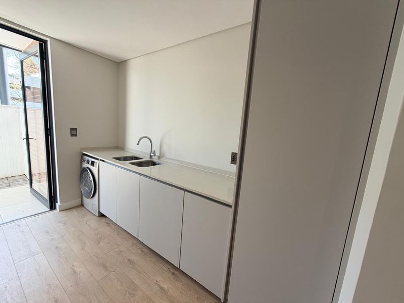 A modern, minimalist laundry room featuring sleek built-in cabinetry, a stone countertop with a stainless steel sink, and an integrated washing machine.