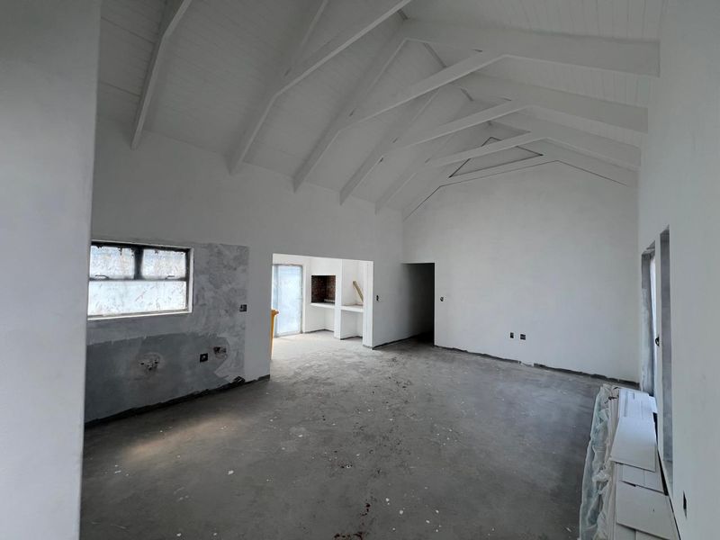 An expansive open-plan living area featuring a high vaulted ceiling with exposed trusses, concrete flooring, and a built-in braai, currently in an unfinished state.