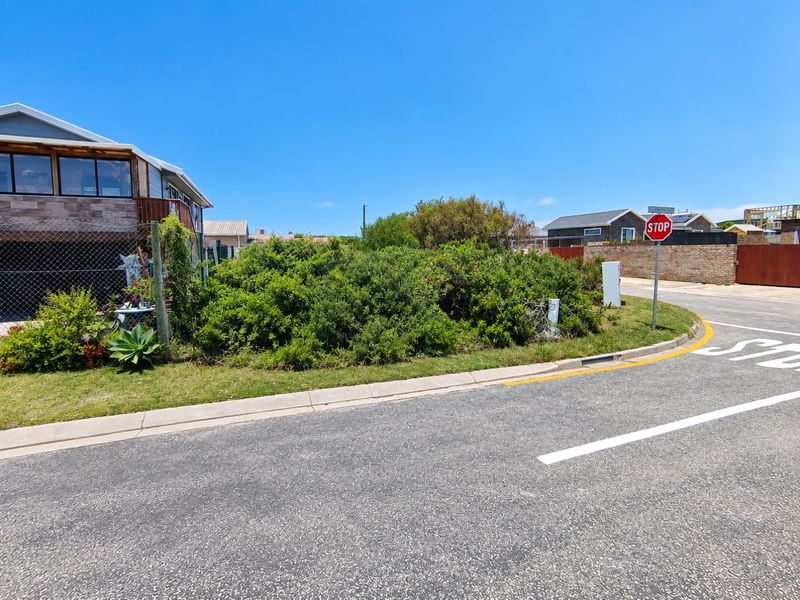 Street view of the vacant residential plot corner, featuring established vegetation, paved road access, and nearby utility infrastructure.