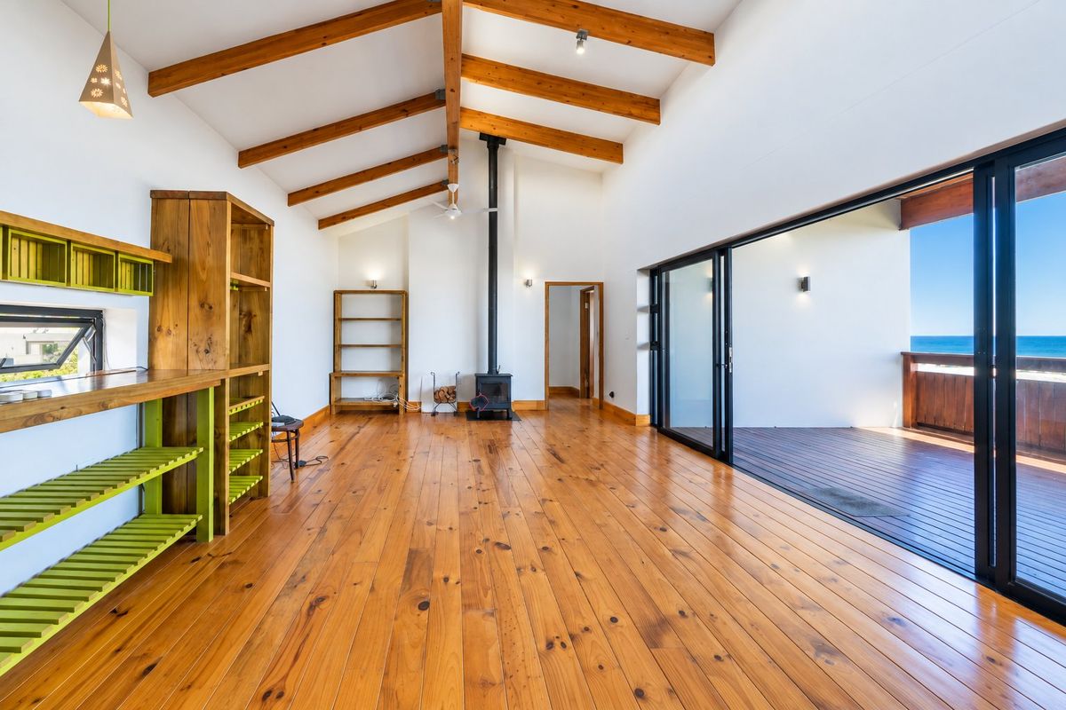 Spacious living area featuring exposed timber beams, hardwood floors, a wood-burning fireplace, and large sliding doors opening to an ocean-view deck.