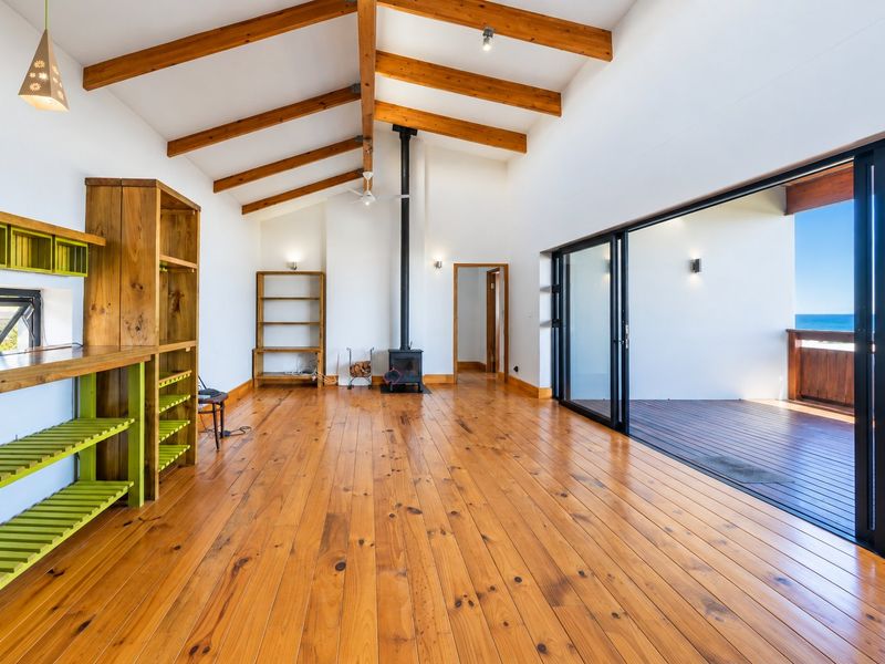 Spacious living area featuring exposed timber beams, hardwood floors, a wood-burning fireplace, and large sliding doors opening to an ocean-view deck.