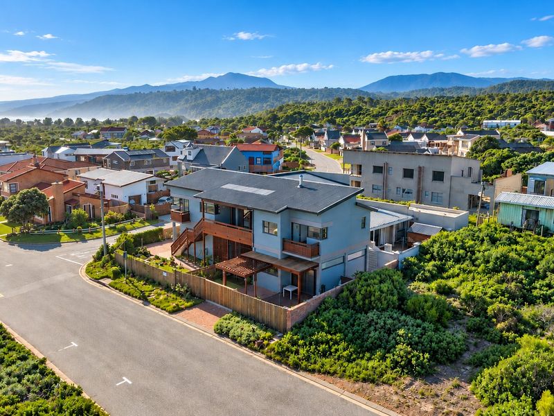 Aerial view of a modern two-story residential property featuring timber decking, a pergola, and solar panels, situated in a coastal neighborhood with mountain views.
