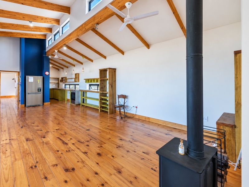 Spacious open-plan living and kitchen area featuring high vaulted ceilings with exposed timber beams, warm wood flooring, and a central wood-burning stove.