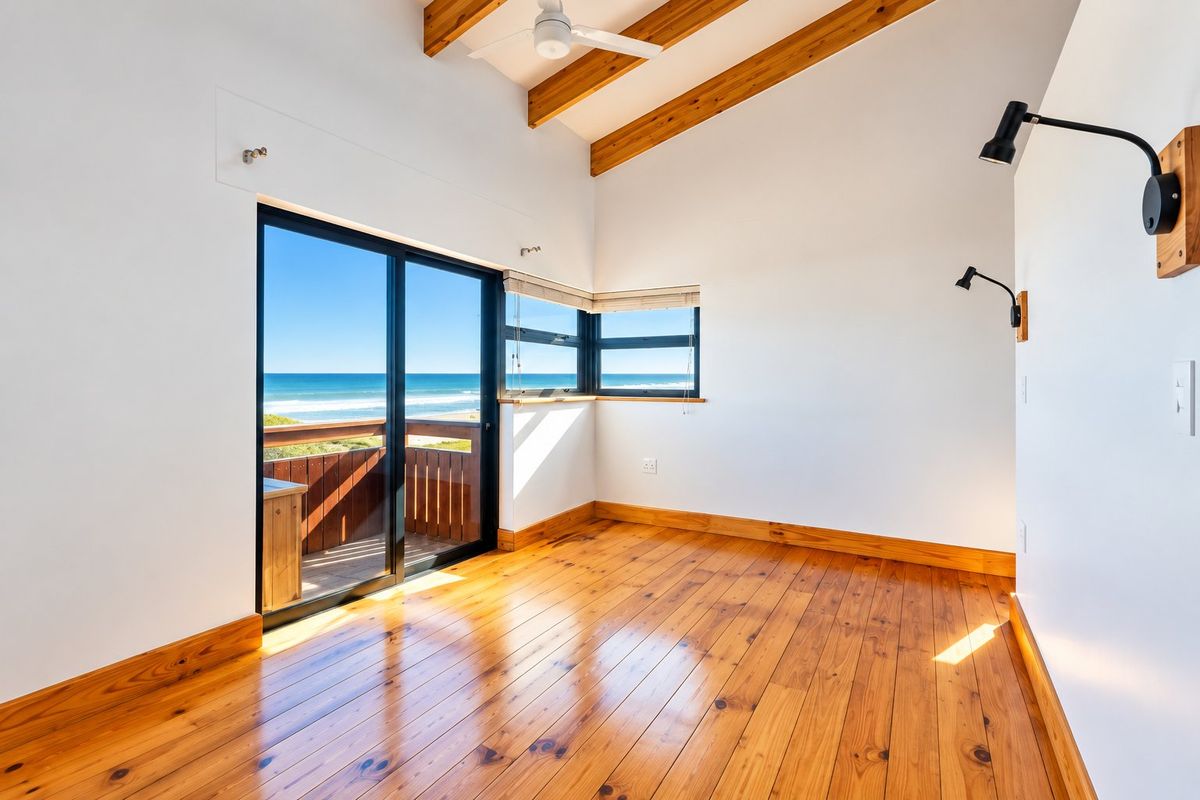 Bright bedroom featuring exposed timber ceiling beams, polished timber flooring, and a sliding glass door opening to a balcony with ocean views.