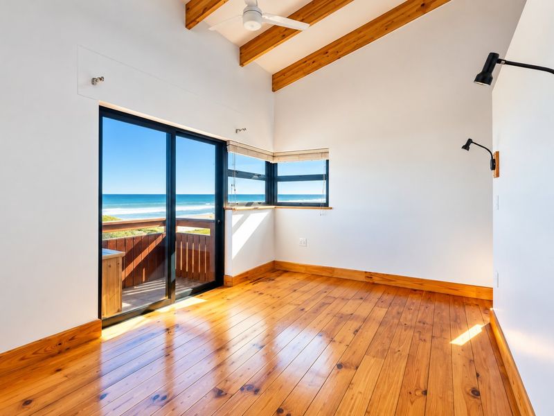 Bright bedroom featuring exposed timber ceiling beams, polished timber flooring, and a sliding glass door opening to a balcony with ocean views.