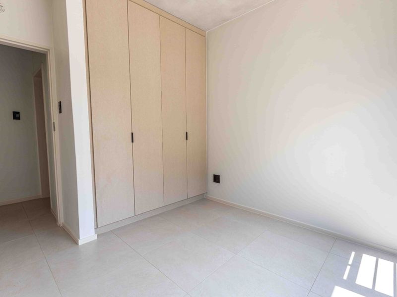 A bright, empty bedroom featuring neutral tiled flooring and a large, modern built-in wardrobe with light wood-finish doors.