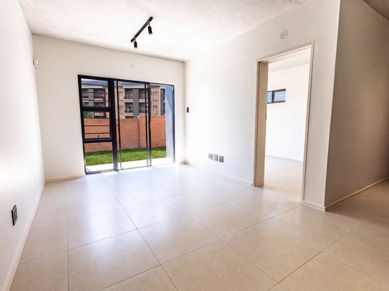 A bright, empty open-plan living area featuring neutral tiled flooring, modern track lighting, and a sliding glass door providing access to the garden.