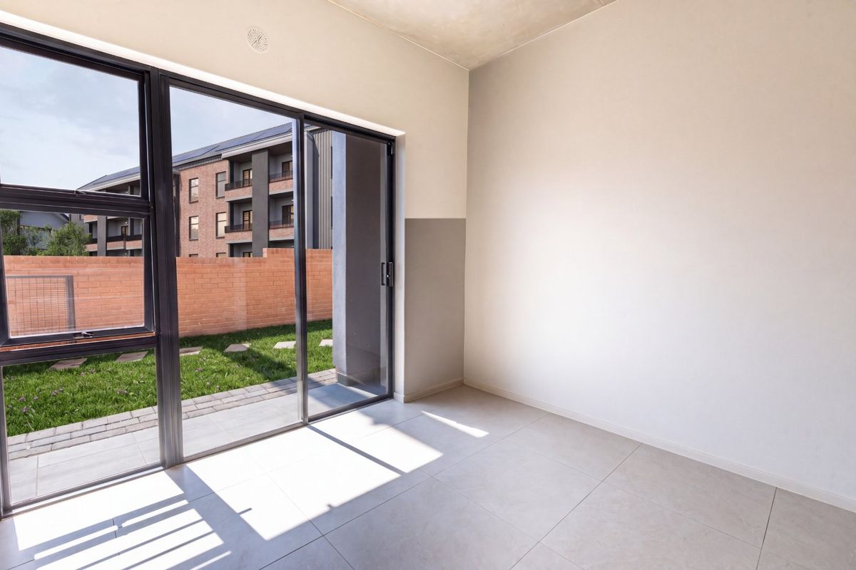 A bright, empty living area featuring large sliding glass doors that open onto a private garden space with modern tiled flooring.