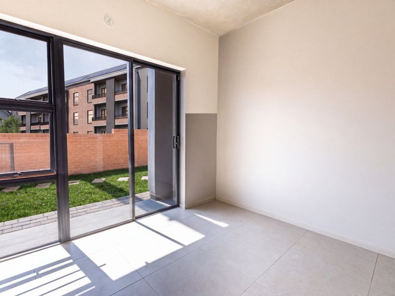 A bright, empty living area featuring large sliding glass doors that open onto a private garden space with modern tiled flooring.