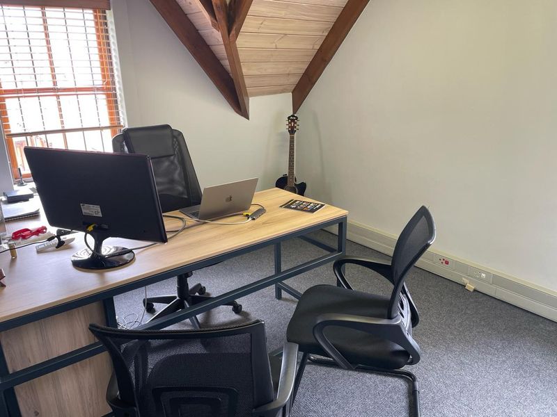 A functional office space featuring exposed timber rafters, carpeted flooring, and natural light from a window with venetian blinds.
