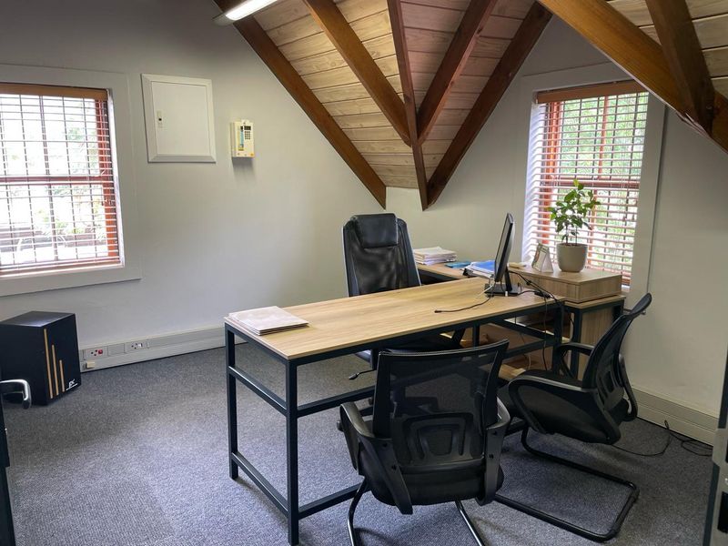 A functional office space featuring exposed timber rafters, carpeted flooring, and natural light from dual windows.