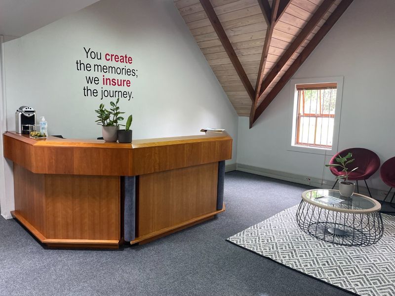 A professional office reception area featuring a built-in wooden desk, exposed timber roof trusses, and a bright window with natural light.