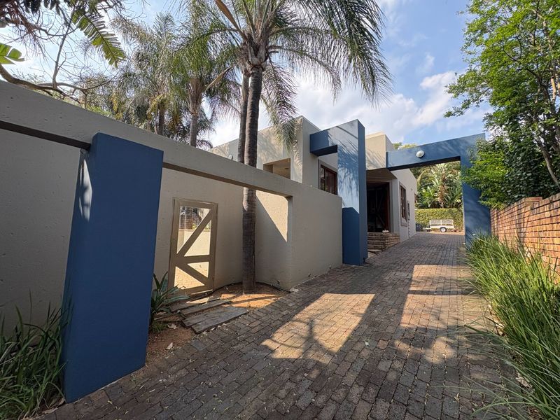 Front exterior view featuring a paved driveway, modern architectural pillars, and a gated entrance with established trees and boundary walls.