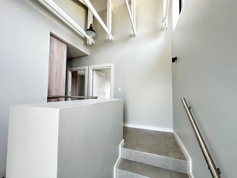 A bright, modern hallway featuring a tiled staircase with a stainless steel handrail, exposed ceiling beams, and clean, neutral finishes.