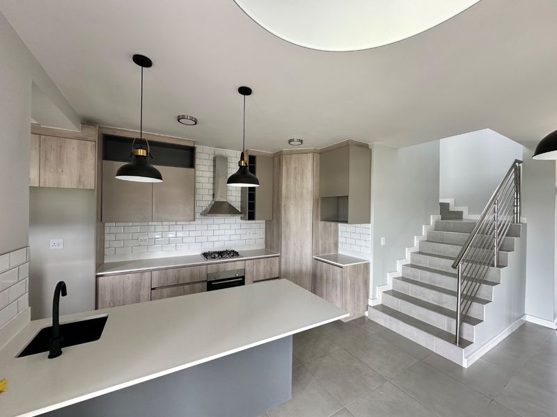 Modern open-plan kitchen featuring light wood cabinetry, stone countertops, subway tile backsplash, and integrated appliances, adjacent to a tiled staircase with stainless steel railing.