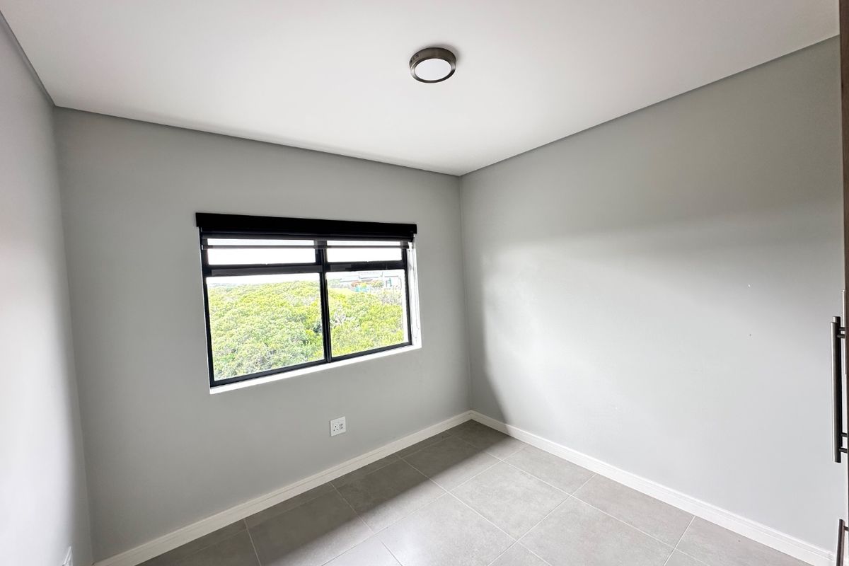 A clean, empty bedroom featuring neutral grey walls, large tiled flooring, and a black-framed window with a roller blind.