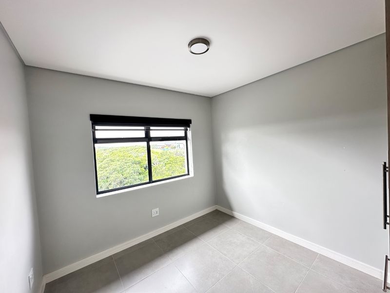 A clean, empty bedroom featuring neutral grey walls, large tiled flooring, and a black-framed window with a roller blind.