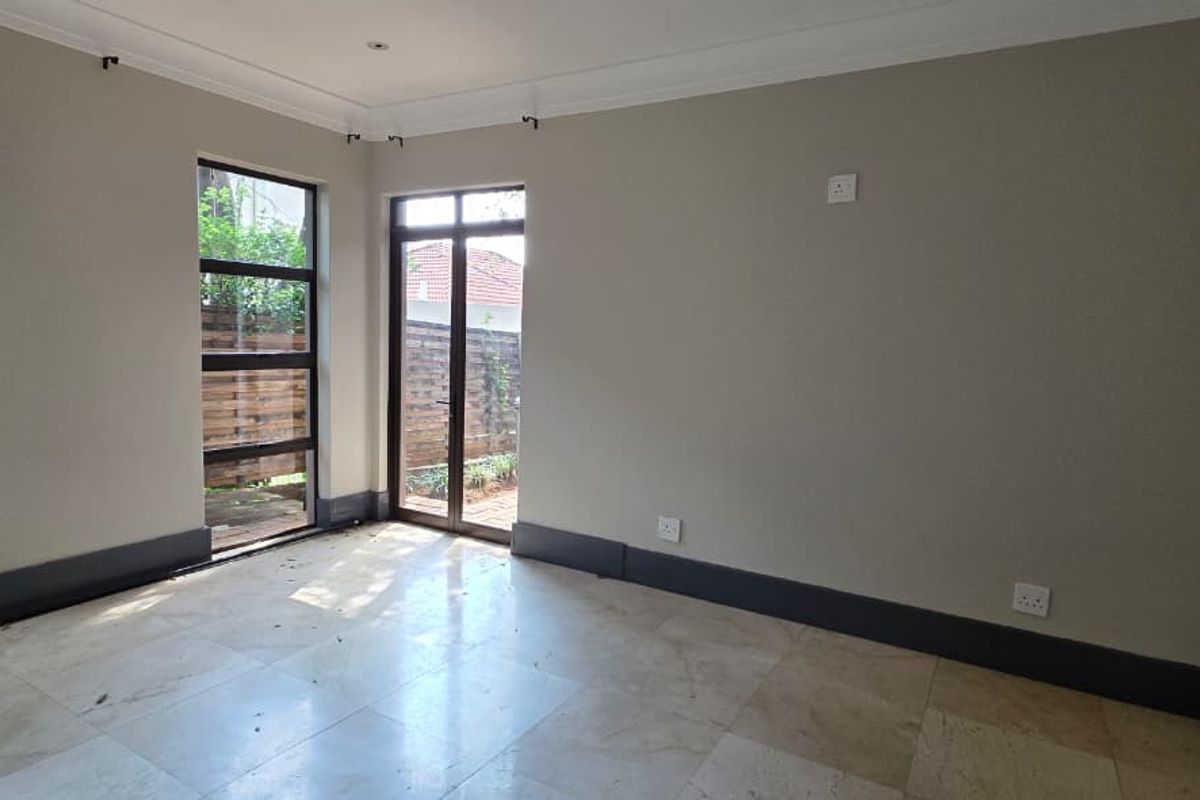 A bright, empty living area featuring neutral walls, stone tile flooring, and large aluminium-framed windows and doors providing access to the garden.