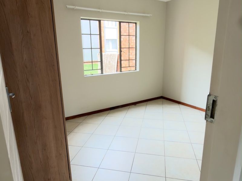 A bright, empty bedroom featuring neutral tiled flooring, a built-in cupboard, and a window with security bars.